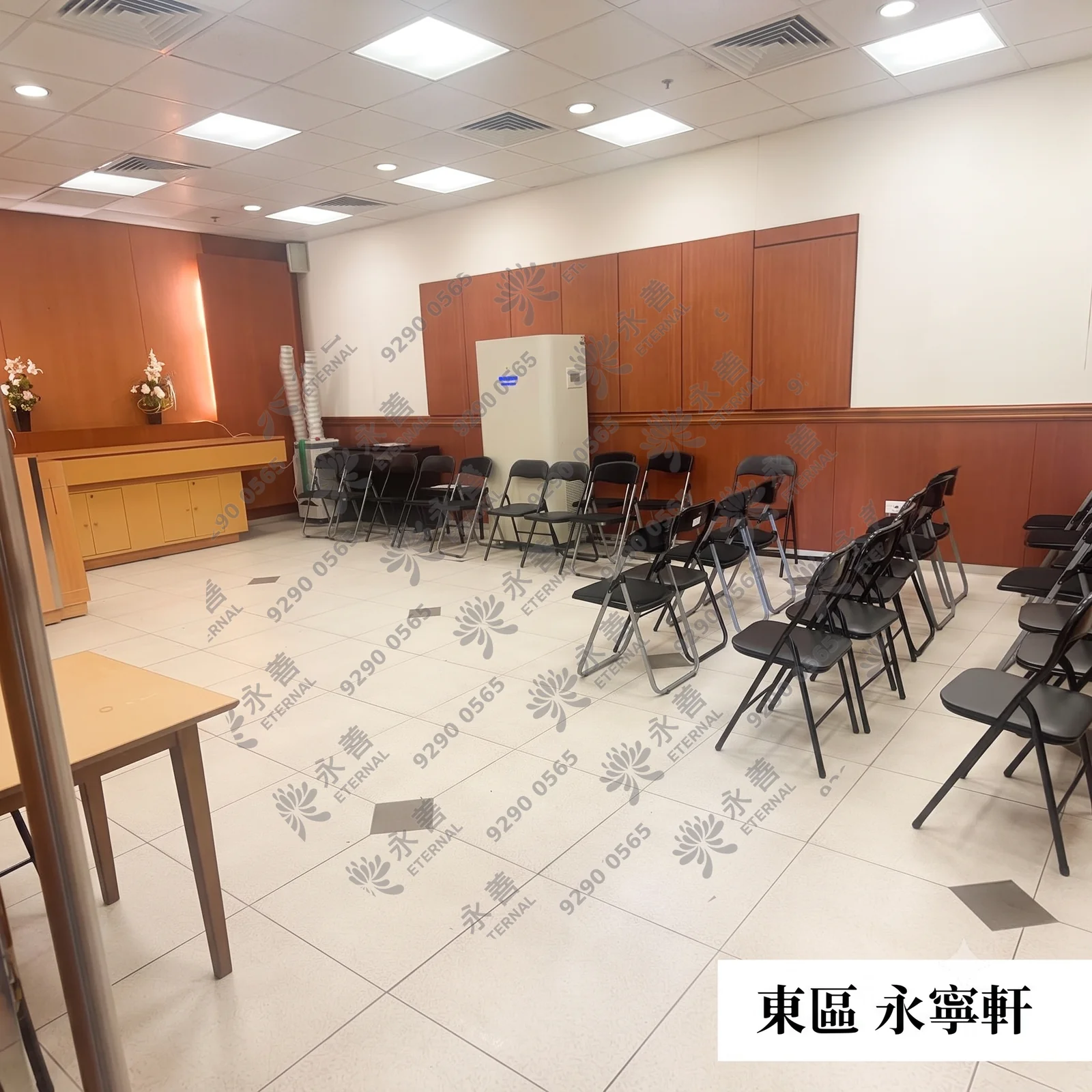 東區醫院永寧軒禮堂內部的實景環境。室內鋪設帶有菱形點綴的淺色地磚，牆面採白色與木質雙色拼接。前方設有木製祭台，台上有鮮花裝飾，場地內整齊排列著多張黑色摺疊椅供親友觀禮。圖片右下角帶有「東區 永寧軒」的白色文字標籤。