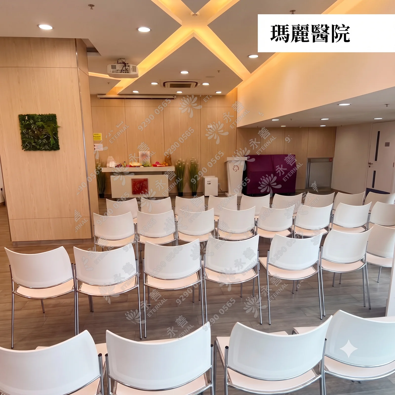 瑪麗醫院告別禮堂的內部實景。室內燈光柔和，天花板有著十字交叉的間接照明設計。前方設有木質講台、祭台及植物造景，台前整齊排列著多排現代風格的白色靠背椅，整體氛圍平靜而莊嚴。右上角附有「瑪麗醫院」的黑體字樣標籤。